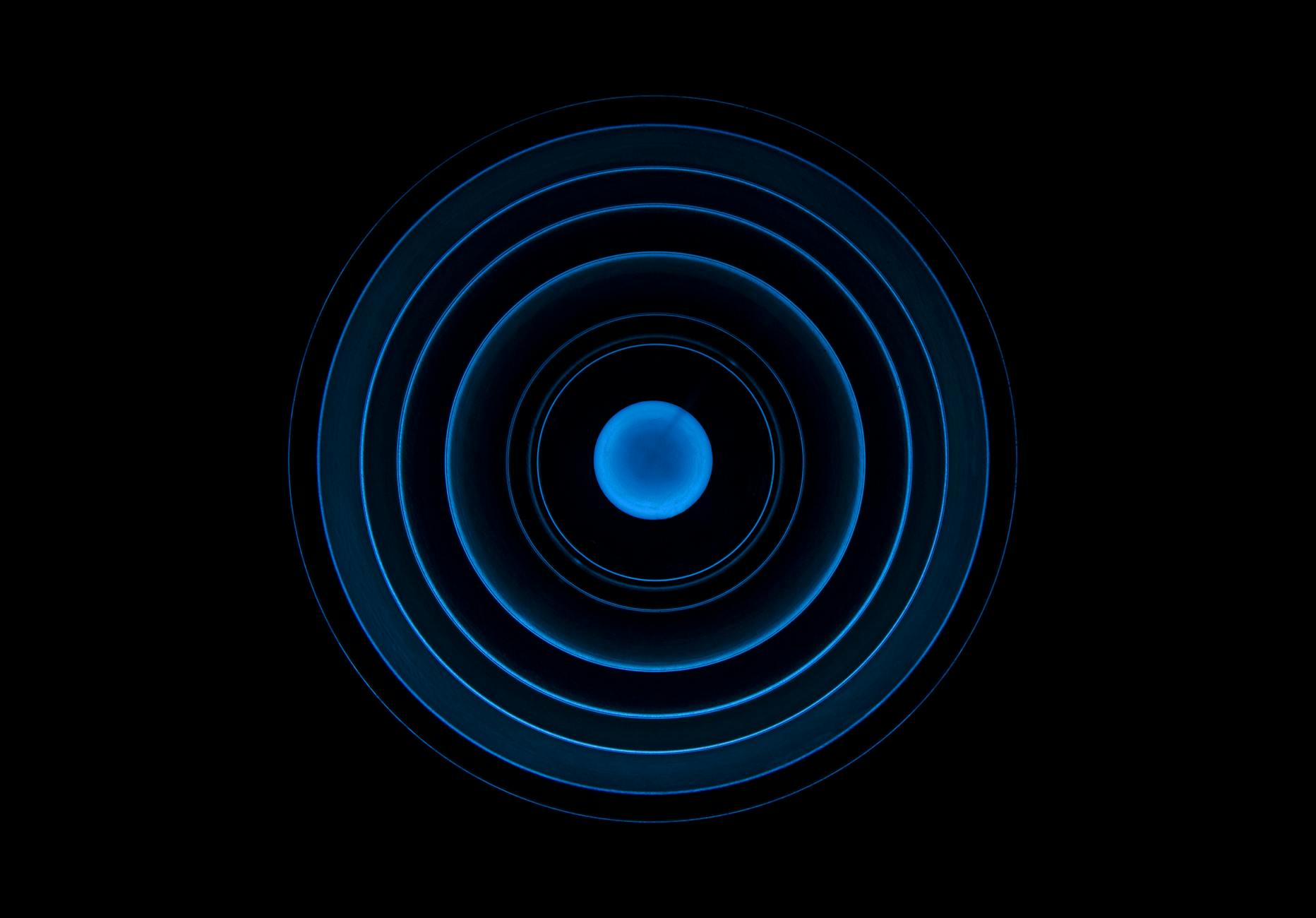A glowing blue circle filled with blue and surrounded by thin circles of blue growing increasingly bigger the further out they go into a black background